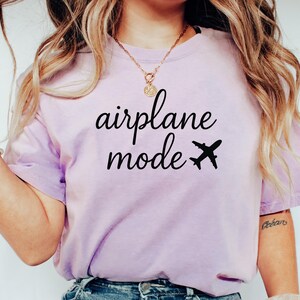 K&ouml;nnte beinhalten: Helllila T-Shirt mit dem schwarzen Schriftzug "airplane mode" und einer schwarzen Flugzeug-Grafik. Das Shirt hat einen l&auml;ssigen Stil.