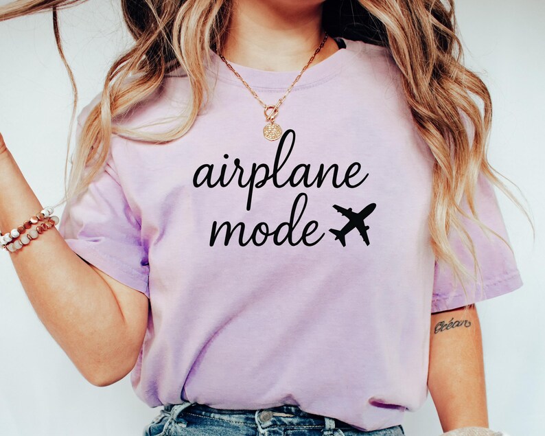 K&ouml;nnte beinhalten: Helllila T-Shirt mit dem schwarzen Schriftzug "airplane mode" und einer schwarzen Flugzeug-Grafik. Das Shirt hat einen l&auml;ssigen Stil.