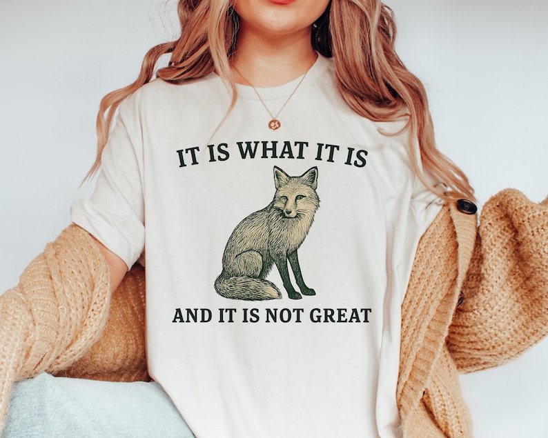Op de afbeelding: Cr&egrave;mekleurig T-shirt met een gedetailleerde illustratie van een vos. De tekst "IT IS WHAT IT IS" staat boven de vos en "AND IT IS NOT GREAT" eronder, allemaal in donkergroene letters. Het shirt heeft korte mouwen.