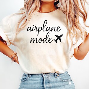 K&ouml;nnte beinhalten: Cremefarbenes T-Shirt mit dem Schriftzug "airplane mode" in schwarzer Schrift, begleitet von einer kleinen Flugzeuggrafik. Das Shirt ist ein l&auml;ssiger Stil, geeignet f&uuml;r Reisen oder den Alltag.