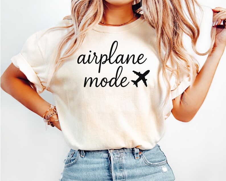 K&ouml;nnte beinhalten: Cremefarbenes T-Shirt mit dem Schriftzug "airplane mode" in schwarzer Schrift, begleitet von einer kleinen Flugzeuggrafik. Das Shirt ist ein l&auml;ssiger Stil, geeignet f&uuml;r Reisen oder den Alltag.