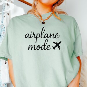 K&ouml;nnte beinhalten: Hellgr&uuml;nes T-Shirt mit dem schwarzen Schriftzug "airplane mode" und einer schwarzen Flugzeug-Grafik. Das Shirt hat einen l&auml;ssigen Stil.