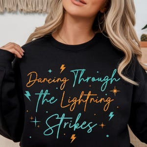 Puede incluir: Sudadera negra con la frase "Dancing Through The Lightning Strikes" en escritura turquesa y naranja. El diseño incluye rayos y estrellas. Una prenda superior casual y cómoda.