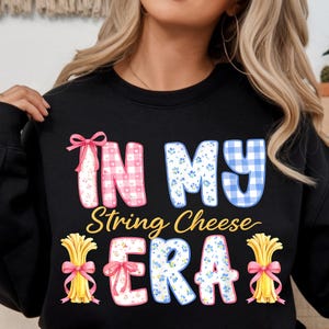 May include: Black sweatshirt featuring the phrase "IN MY String Cheese ERA". The text uses pink gingham, blue floral patterns, and yellow string cheese bundles tied with pink bows. A playful, food-related design.