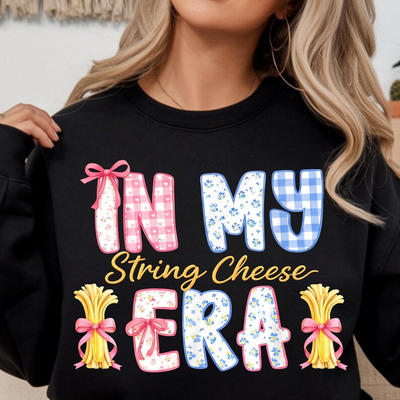 May include: Black sweatshirt featuring the phrase "IN MY String Cheese ERA". The text uses pink gingham, blue floral patterns, and yellow string cheese bundles tied with pink bows. A playful, food-related design.
