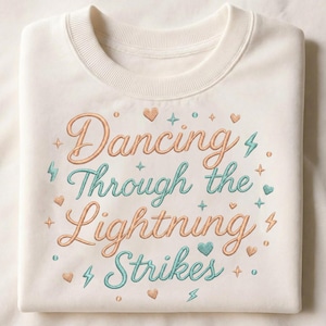 May include: Cream-colored sweatshirt with the embroidered phrase "Dancing Through the Lightning Strikes" in peach and teal. The design includes hearts, stars, and lightning bolt accents, creating a whimsical and stylish look.