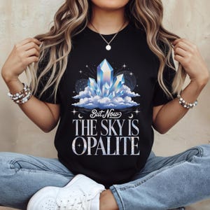 May include: Black t-shirt featuring a crystal design with blue, white, and purple hues, set against a backdrop of clouds and stars. The text "But Now The Sky Is Opalite" is displayed below the graphic.