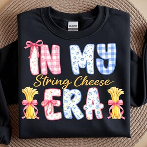 May include: Black sweatshirt featuring the text "IN MY String Cheese ERA". The text is in a variety of patterns, including gingham and floral. The design includes illustrations of string cheese bundles tied with pink bows.
