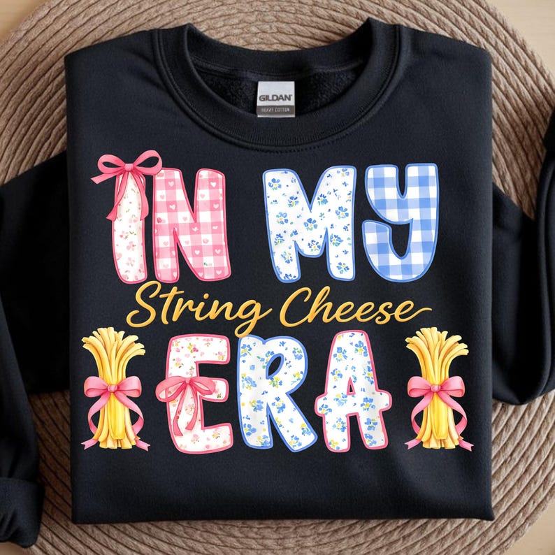 May include: Black sweatshirt featuring the text "IN MY String Cheese ERA". The text is in a variety of patterns, including gingham and floral. The design includes illustrations of string cheese bundles tied with pink bows.