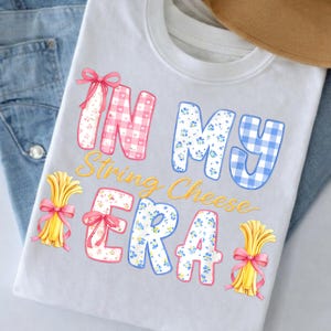 May include: White t-shirt featuring the text "IN MY String Cheese ERA" in a playful, colourful design. The letters are decorated with pink gingham, blue floral, and blue gingham patterns, alongside illustrations of wheat bundles tied with pink bows.