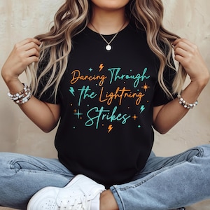 Puede incluir: Camiseta negra con la frase "Dancing Through The Lightning Strikes" en naranja y turquesa. El diseño incluye rayos y estrellas. Prenda de vestir informal.