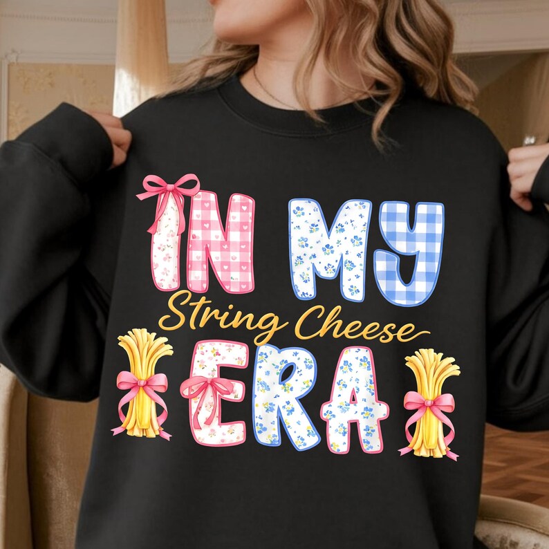 May include: Black sweatshirt featuring the phrase "IN MY String Cheese ERA". The text uses a whimsical font and is decorated with gingham, floral, and plaid patterns. The design includes string cheese illustrations.