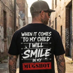 May include: A black short-sleeved t-shirt featuring white text that reads: WHEN IT COMES TO MY CHILD I WILL SMILE IN MY MUGSHOT, set against a background of horizontal height measurement lines and a red rectangular box around the word MUGSHOT.