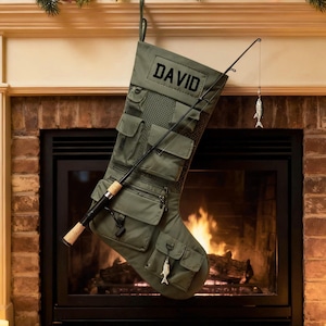 May include: An olive green Christmas stocking with multiple pockets and the name "DAVID" printed on it. A fishing rod and a small fish ornament are attached. The stocking hangs in front of a fireplace.