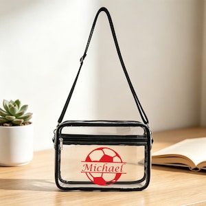 Peut inclure: Un sac bandoulière transparent avec une sangle et des bordures noires. Le sac présente un graphisme de ballon de football rouge avec le nom "Michael" en rouge. Le sac est rectangulaire avec une fermeture éclair.