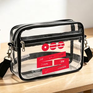 May include: A clear, rectangular crossbody bag with black trim and a black adjustable shoulder strap. The bag features red accents with text and a logo. The bag is sitting on a wooden surface.