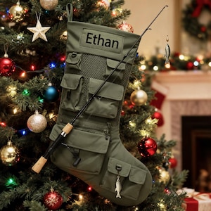 Peut inclure: Chaussette de Noël vert olive avec plusieurs poches, une canne à pêche et un charme en forme de poisson. Le nom "Ethan" est imprimé sur la chaussette. La chaussette est décorée d'une canne à pêche et d'un charme en forme de poisson. La chaussette est accrochée à un sapin de Noël.
