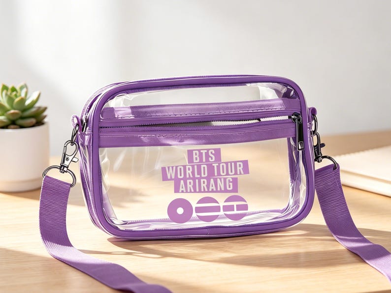May include: A clear, rectangular crossbody bag with a purple trim, zipper, and strap. The bag features the text "BTS WORLD TOUR ARIRANG" in white. The bag is sitting on a wooden surface.