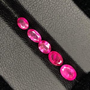 以下が含まれることがあります： 斜めのラインに並べられた、5つのオーバルカットの鮮やかなピンクがかった赤い宝石。それぞれの石は光を反射するファセット加工された表面を持ち、輝きのある半透明の品質を示しています。宝石は暗い質感のある表面に一列に配置されています。