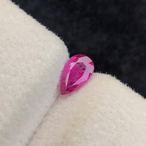 May include: A faceted, pear-shaped, vibrant pink gemstone. The gem is displayed on a white fabric against a dark background. The cut of the stone allows light to reflect, creating a sparkling effect. This gemstone is likely for jewelry making.