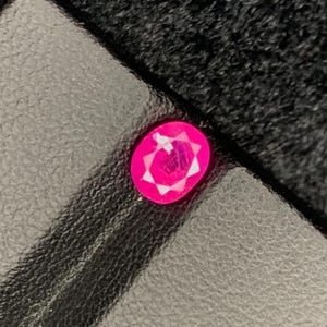 Könnte beinhalten: Ein facettierter, ovalförmiger, leuchtend rosa Edelstein liegt auf einer schwarzen, lederartigen Oberfläche. Der Schliff des Edelsteins zeigt mehrere Facetten, die einen Sterneneffekt erzeugen. Der Hintergrund ist ein dunkles, strukturiertes Gewebe.