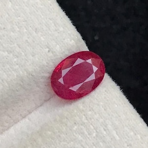 Puede incluir: Una gema de rubí facetada de forma ovalada. La gema es de color rojo intenso y está colocada sobre una superficie blanca con un fondo negro. Las facetas del rubí crean un patrón de estrella.
