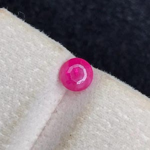 Puede incluir: Una pequeña piedra preciosa redonda y facetada con un tono rosa rojizo vibrante y saturado. La piedra está colocada en el borde de una tarjeta de presentación rectangular de color blanco roto y texturizada, mostrando su superficie pulida.