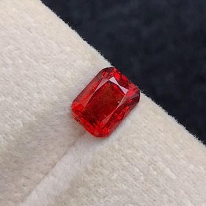 以下が含まれることがあります： 鮮やかな長方形カットの赤い宝石。宝石は、暗い背景に対して、白くテクスチャ加工された表面に表示されています。宝石のファセットは光を反射し、その透明度と色を示しています。