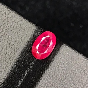 Könnte beinhalten: Ein oval geschliffener Rubin-Edelstein in leuchtendem Rot. Der Edelstein liegt auf einer strukturierten, dunkelgrauen Oberfläche, was einen starken Kontrast erzeugt. Die Facetten des Rubins fangen das Licht ein und zeigen seinen Glanz.