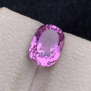 Puede incluir: Una gema pulida, de corte ovalado, en tonos rosa y lavanda. La gema está colocada sobre una superficie blanca, con un fondo oscuro. Las facetas de la gema reflejan la luz, creando un efecto brillante. Esta gema es adecuada para joyería.