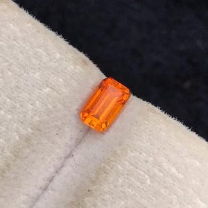 Pode incluir: Uma gema de cor laranja vibrante cortada em formato retangular de esmeralda. A pedra apresenta facetas claras que refletem a luz, criando uma aparência brilhante. Está posicionada sobre uma superfície clara e texturizada com aspeto de feltro.