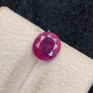 May include: A loose oval-cut ruby gemstone with a vibrant deep pinkish-red hue and a faceted surface that reflects light. The gemstone is positioned on a light-colored, textured display card that features a vertical crease down the center.