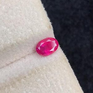 May include: A vibrant, oval-cut pink gemstone with a faceted surface, resting on a textured, off-white fabric display stand. The gemstone exhibits a brilliant, reflective quality, showcasing its clear, polished structure against the neutral material.