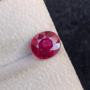 May include: A loose, oval-cut ruby gemstone with a deep red color and faceted surface. The gemstone is positioned on a textured, off-white display card, showcasing its brilliant clarity and geometric cut from a top-down perspective.