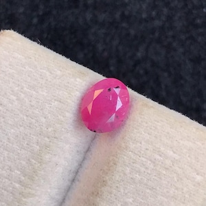 May include: A vibrant, oval-cut pink gemstone with multiple facets reflecting light, resting on a textured, off-white display stand. The stone exhibits a deep magenta hue and a polished surface, showcasing its clear, geometric cut and natural clarity.