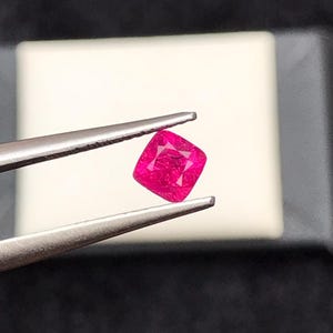 Peut inclure: Un rubis carré, de couleur rose-rouge vif, maintenu par une pince argentée. La pierre précieuse présente une surface légèrement texturée. L'arrière-plan est blanc et gris neutre.