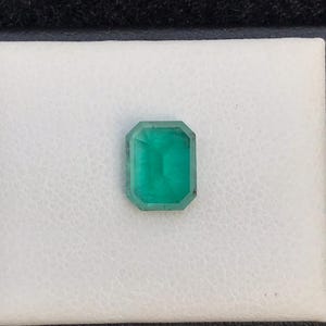 Puede incluir: Una piedra preciosa de esmeralda rectangular con un corte facetado. La gema es de color verde vibrante y est&aacute; colocada sobre un fondo blanco. La esmeralda es una piedra preciosa, a menudo utilizada en joyer&iacute;a.