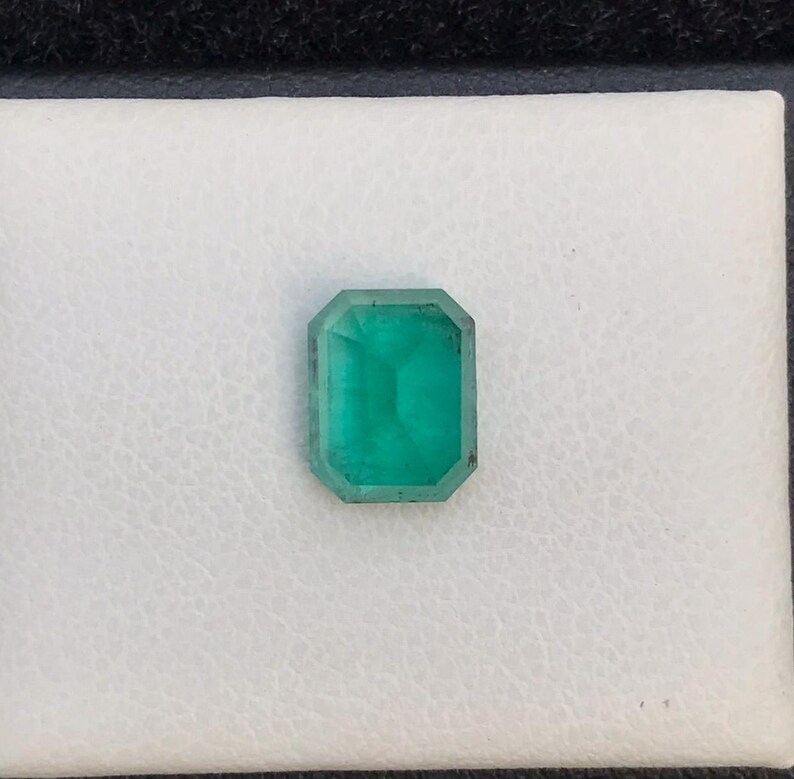 Puede incluir: Una piedra preciosa de esmeralda rectangular con un corte facetado. La gema es de color verde vibrante y est&aacute; colocada sobre un fondo blanco. La esmeralda es una piedra preciosa, a menudo utilizada en joyer&iacute;a.