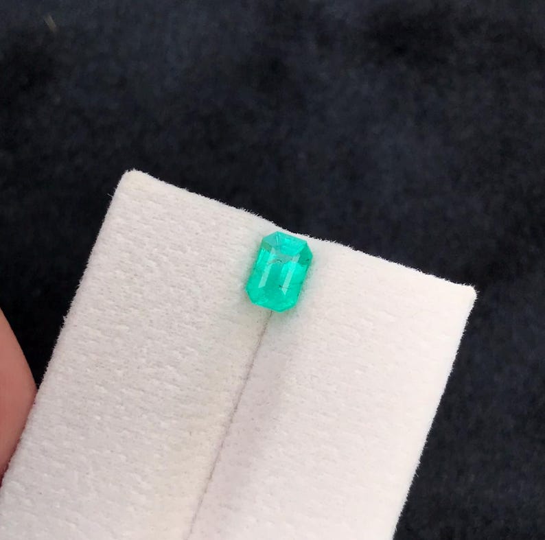 Puede incluir: Una esmeralda rectangular verde brillante descansa sobre una superficie blanca. Las facetas de la gema reflejan la luz, mostrando su claridad y corte. El fondo es de un color oscuro, proporcionando contraste.