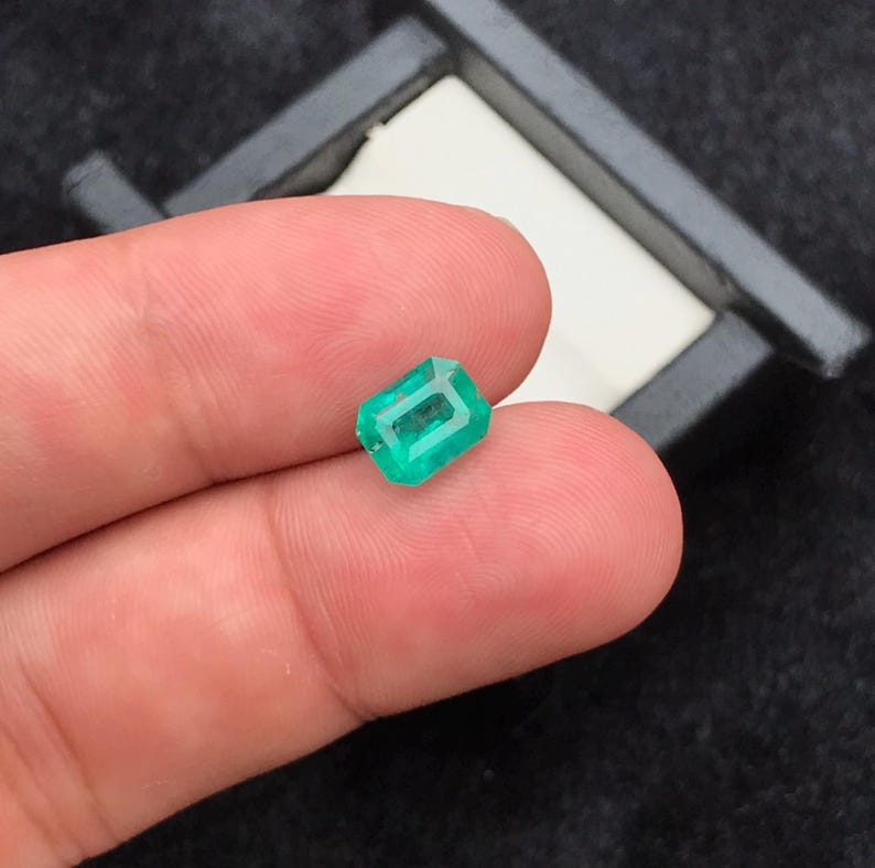 Puede incluir: Una esmeralda rectangular verde brillante. La gema se sostiene entre los dedos, mostrando su claridad y corte. La esmeralda se coloca sobre un fondo neutro, resaltando su color y brillo. La piedra preciosa es una piedra preciosa.
