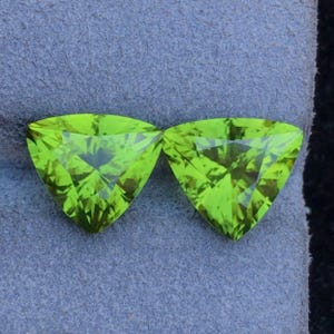 Op de afbeelding: Twee groene peridot edelstenen, geslepen in een driehoekige vorm, met een gefacetteerde afwerking. De edelstenen zijn geplaatst tegen een blauwe fluwelen achtergrond.