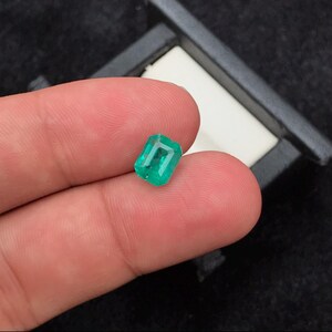 Puede incluir: Una esmeralda de corte rectangular, de color verde vibrante. La gema se muestra sobre un fondo oscuro, sostenida entre los dedos. El corte esmeralda resalta la claridad y el color de la piedra.