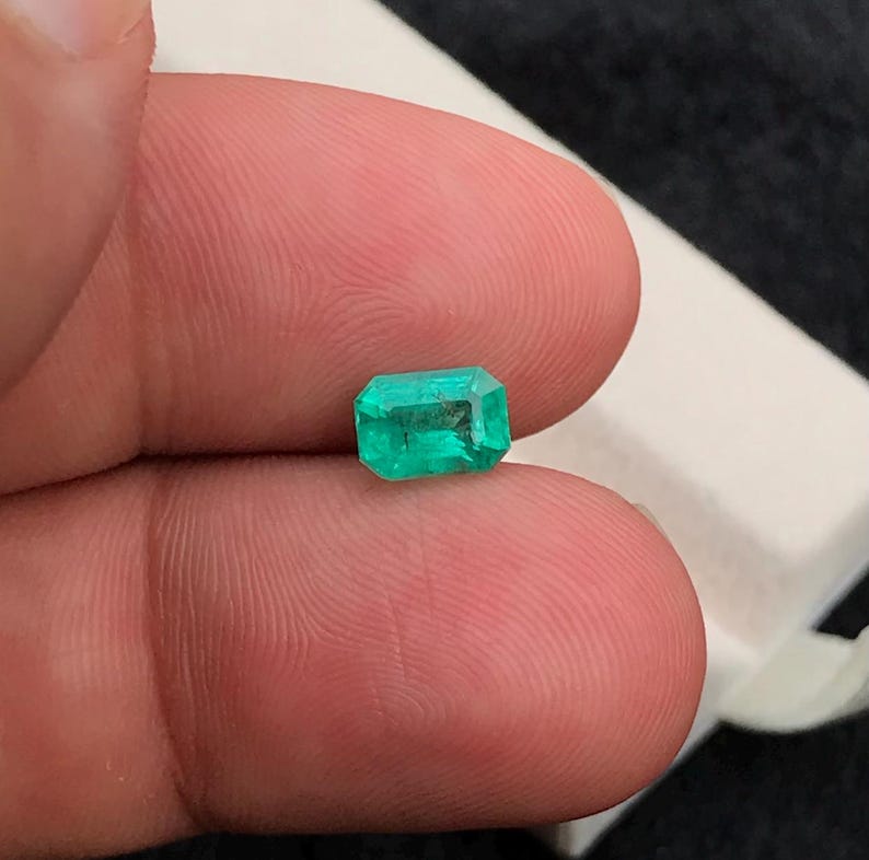 Puede incluir: Una esmeralda rectangular de color verde brillante. La gema se sostiene entre los dedos. La esmeralda tiene una superficie clara y pulida, mostrando sus facetas. La piedra preciosa es un punto focal, lo que sugiere joyer&iacute;a o un art&iacute;culo de colecci&oacute;n. Medidas en mil&iacute;metros.