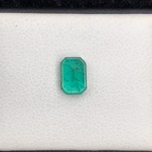 Puede incluir: Una esmeralda de corte rectangular, de color verde vibrante, se muestra sobre un fondo blanco texturizado. La gema est&aacute; engastada en un marco negro, resaltando sus facetas y claridad. Esta gema es ideal para la fabricaci&oacute;n de joyas.