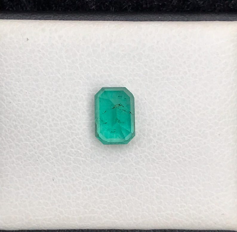 Puede incluir: Una esmeralda de corte rectangular, de color verde vibrante, se muestra sobre un fondo blanco texturizado. La gema est&aacute; engastada en un marco negro, resaltando sus facetas y claridad. Esta gema es ideal para la fabricaci&oacute;n de joyas.