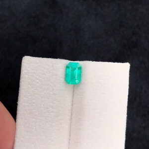 Puede incluir: Una esmeralda verde vibrante, tallada en forma rectangular. La gema se muestra sobre un fondo blanco, resaltando su claridad y color. La esmeralda es una joya brillante y llamativa.
