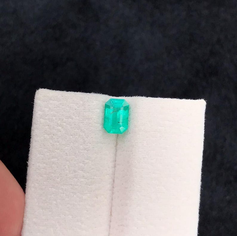 Puede incluir: Una esmeralda verde vibrante, tallada en forma rectangular. La gema se muestra sobre un fondo blanco, resaltando su claridad y color. La esmeralda es una joya brillante y llamativa.