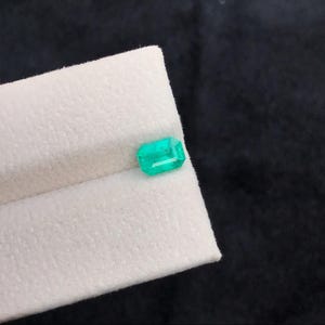 Puede incluir: Una esmeralda rectangular verde brillante. La gema se exhibe sobre una superficie blanca y texturizada contra un fondo oscuro. Las facetas de la esmeralda reflejan la luz, resaltando su claridad y corte. Una hermosa piedra preciosa.
