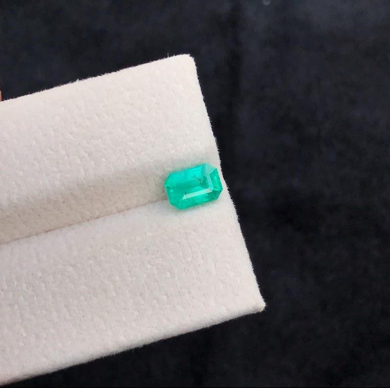Puede incluir: Una esmeralda rectangular verde brillante. La gema se exhibe sobre una superficie blanca y texturizada contra un fondo oscuro. Las facetas de la esmeralda reflejan la luz, resaltando su claridad y corte. Una hermosa piedra preciosa.