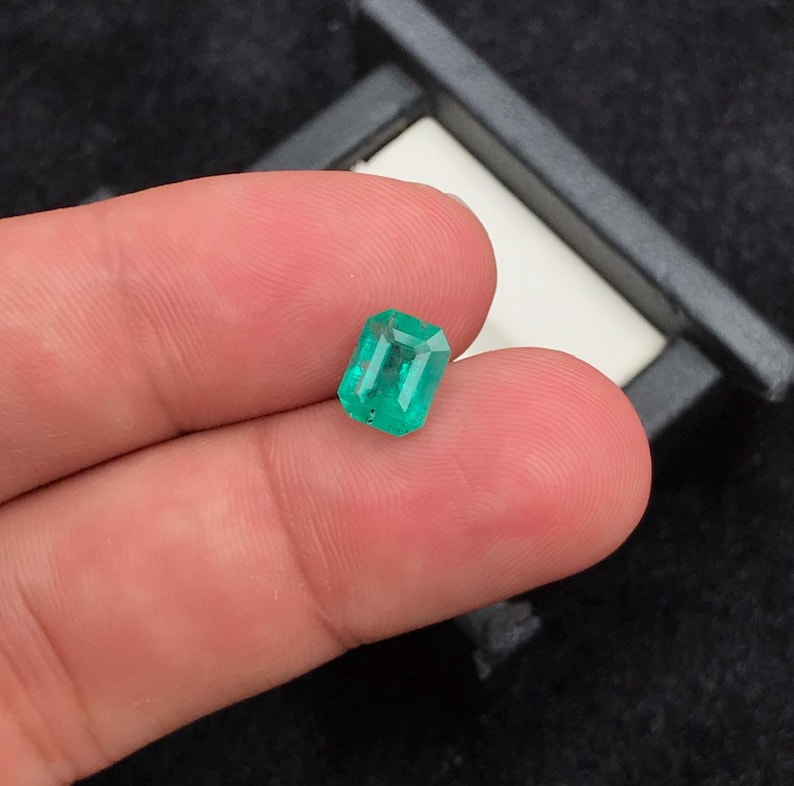 Puede incluir: Una gema de esmeralda rectangular con un tono verde vibrante. La gema se sostiene entre los dedos, mostrando sus facetas y claridad. La esmeralda se encuentra sobre un fondo oscuro, resaltando su color y brillo.